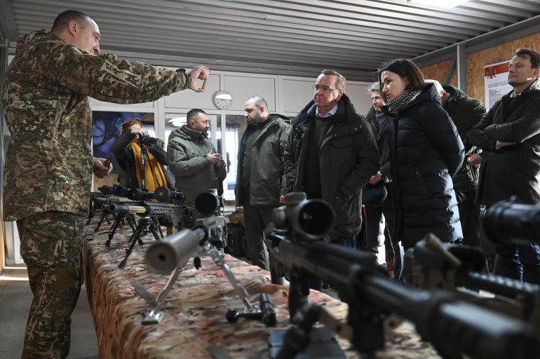 Menteri Pertahanan Jerman Boris Pistorius mendengarkan penjelasan tentang senjata serbu selama kunjungan mereka ke fasilitas pelatihan di luar Kyiv.  Ada banyak senjata yang dipajang di atas meja, dan seorang prajurit menjelaskan masing-masing senjata. 
