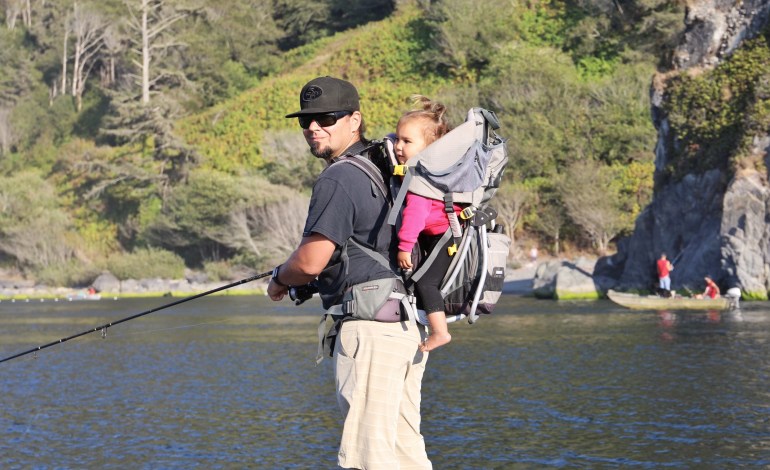 A man, Barry McCovey, stands on the banks of the Klamath River with a fishing pole in hand, as he looks over his shoulder at the camera and reels in the line. On his back is his young child, in a baby carrier.