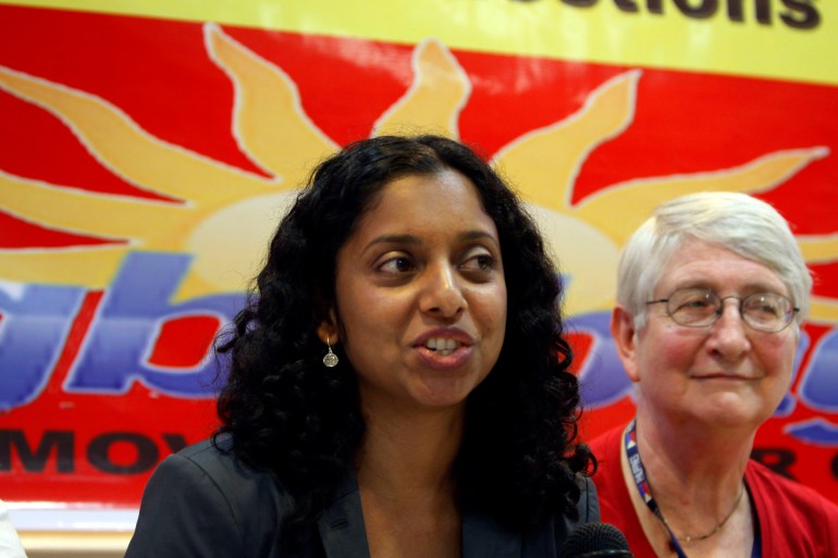 A woman with dark curly hair speaks as she stands or sits next to another woman, with short, white hair. Behind them is a banner with a yellow sun.