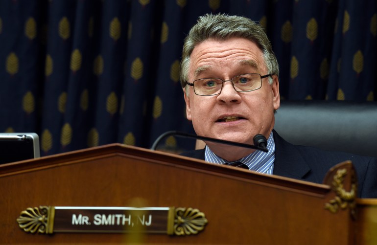Rep. Chris Smith sits behind a wooden sign with his name and the state of New Jersey on it.  He wears glasses and speaks into a microphone.