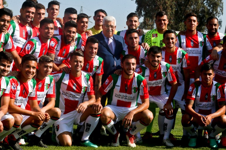 A football team poses in two lines: one kneeling, one standing.  They are members of Club Deportivo Palestino and their jerseys feature the red, green and white stripes of the Palestinian flag.