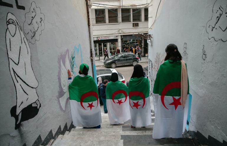young Algerian women wrapped in Algerian flags pose next to street art