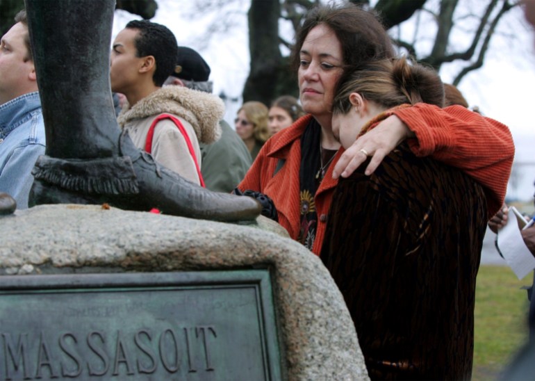 A woman closes her eyes and wraps her arms around another woman, who leans into her comrade, seemingly overcome with emotion. In the foreground, the feet of a bronze statue of Massasoit are visible.