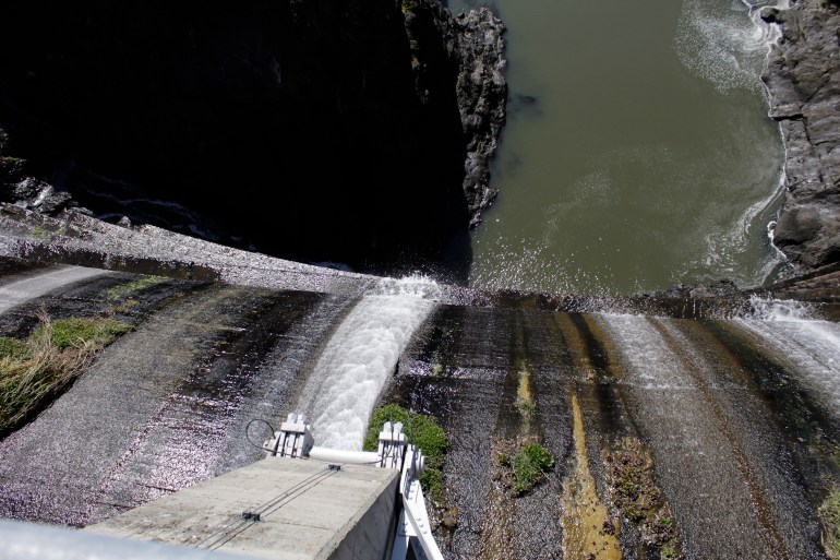 Uma vista do topo de uma barragem, enquanto a água flui pelas curvas de concreto até o rio abaixo.