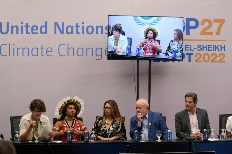 Indigenous leader Puyr Tembe, wearing a crown of radiating feathers, sits on a United Nations Climate Change panel with Brazilian President Lula da Silva, who is dressed in a blue suit. A TV screen broadcasts their remarks above them. 