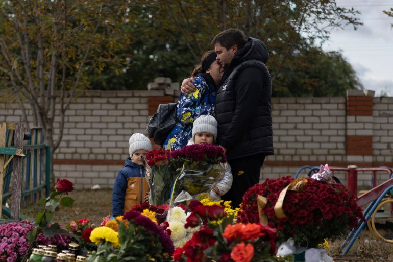 A family visit a memorial for those killed in Hroza. There are lots of flowers. The parents are hugging each other