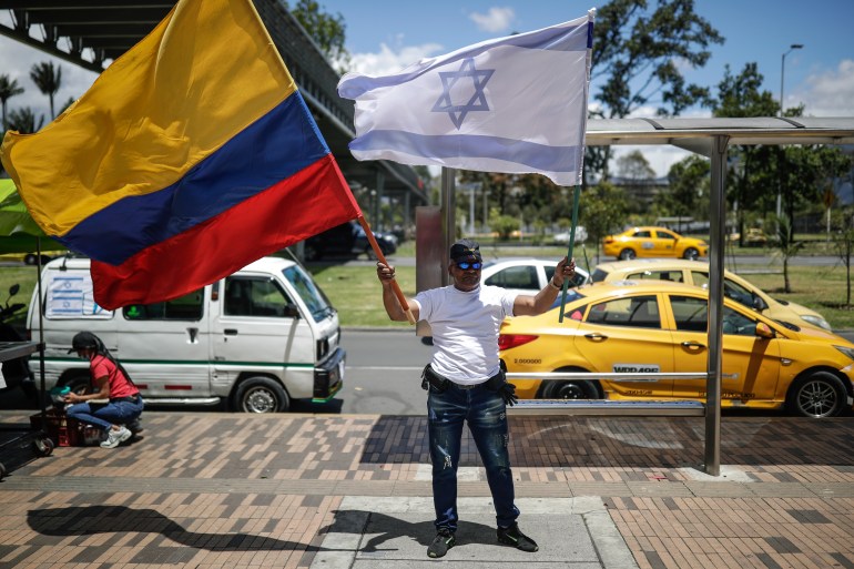 A protester holds up the Colombian flag in one hand and the Israeli flag in the other. He stands on a sidewalk, in front of taxis and a white van passing on the street.
