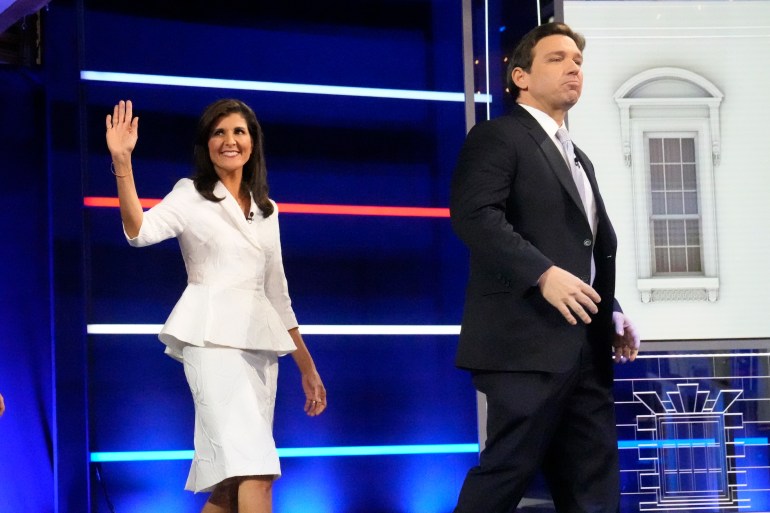 Ron DeSantis entra no palco do debate da NBC em Miami, vestindo terno escuro e gravata clara.  Atrás dele, Nikki Haley segue, acenando para a multidão e vestindo um terno branco.
