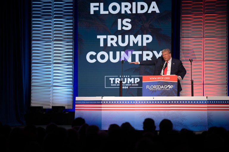 Donald Trump, de terno escuro e gravata vermelha, aponta atrás de um pódio em um comício de campanha.  Atrás dele há uma placa que diz: Flórida é o país de Trump.