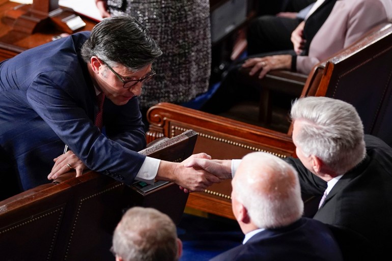Mike Johnson ubrany w ciemny garnitur pochyla się przez oparcie swojego drewnianego krzesła kongresowego, aby uścisnąć dłoń Kevinowi McCarthy’emu.
