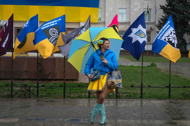 A woman in Kherson stands in front of Ukrainian flags wearing an outfit in the colours of Ukraine beneath a blue and yellow umbrella.