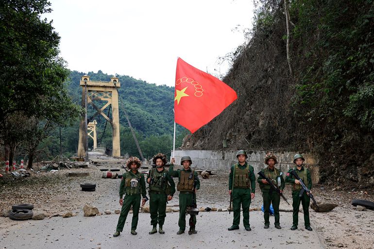 Fighters from the Myanmar National Democratic Alliance Army pose for a photo hold their flag in front of the Kunlong bridge. There are trees on either side of the road leading up to the bridge.