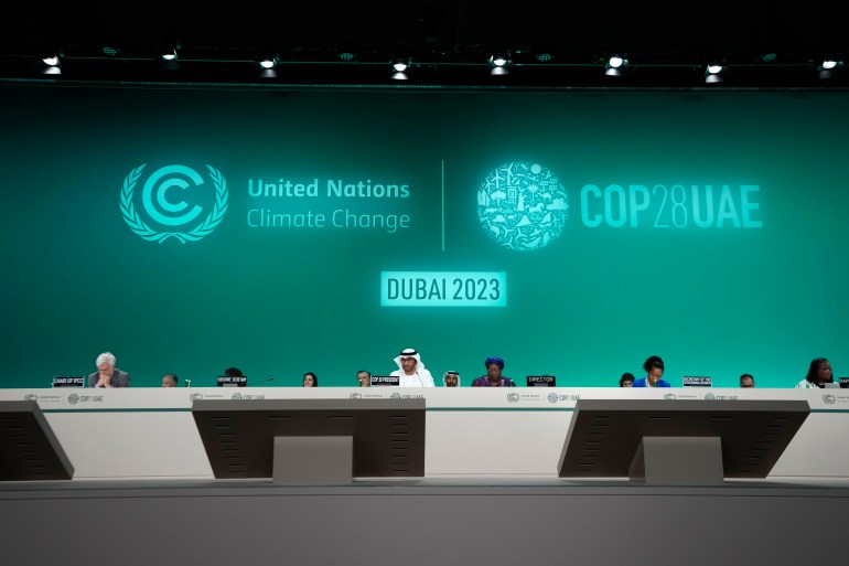 A panel table sits along the length of a stage in Dubai, behind which officials sit. The background of the stage is a green screen with logos like "Dubai 2023" and "COP UAE."