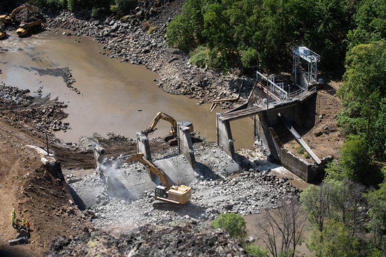 Escavadeiras pesadas derrubam as paredes de concreto de uma barragem no rio Klamath, vista de cima em meio a uma paisagem montanhosa coberta de árvores e arbustos.