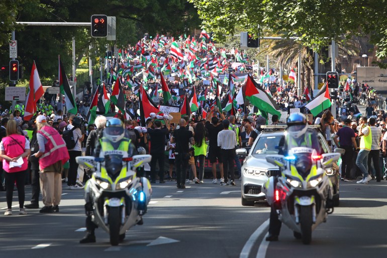 police motorbikes can be seen in front of a large crowd waving Palestinian flags on a sunny day