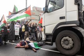 people lie in front of a truck with the Palestinian flag and shipping containers in the background