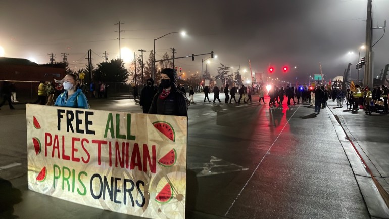 Protesters on a rain-slick road hold up a banner that reads, "Free All Palestinian Prisoners", decorated in watermelon slices to symbolize the colors of the Palestinian flag.
