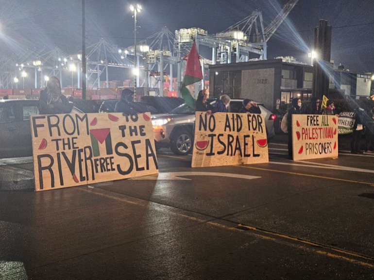 Protesters form a line across a rain-slick road leading to a port. They hold up three large banners. 