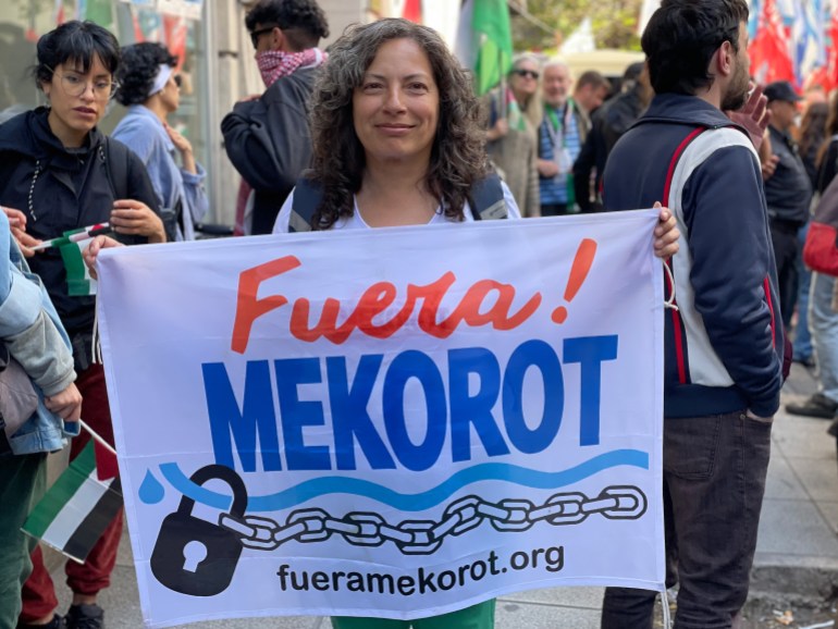 A woman holds up a white banner reading "Fuera! Mekorot," with a wavy line depicting water and an illustration of a chain printed across it.