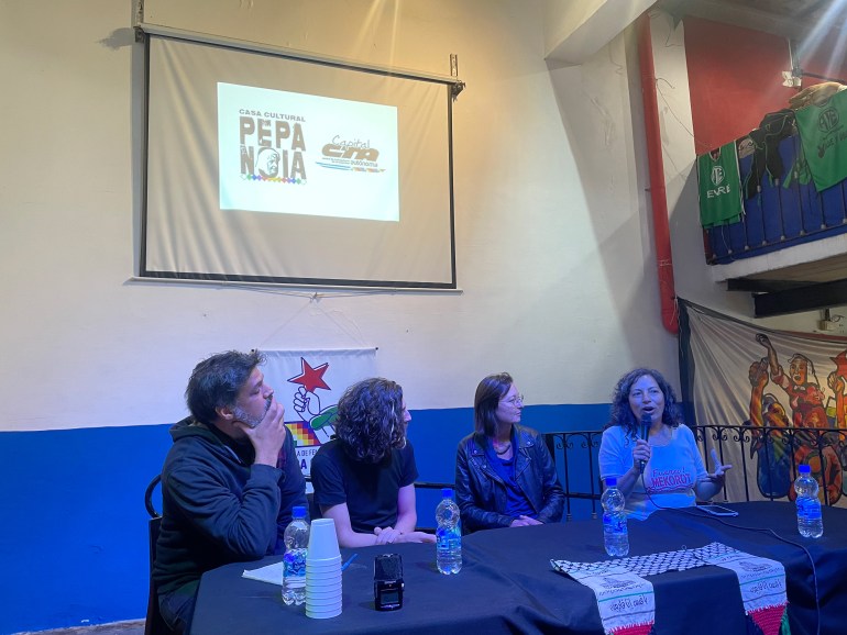 A panel of speakers sits at a table, where Silvia Ferreyra speaks into a microphone. Behind them, a projection on a screen mounted on the wall indicates they are in the Casa Cultural Pepa Noia.