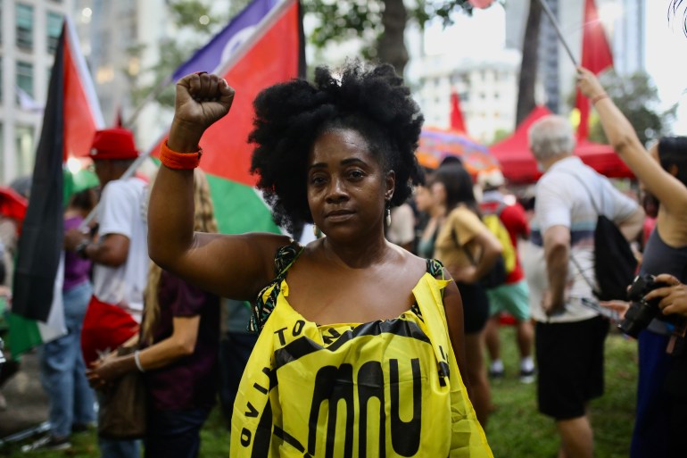 Seorang wanita yang mengenakan spanduk kuning yang diselipkan di bagian depan kemejanya mengangkat tangan sebagai bentuk solidaritas pada protes pro-Palestina di Rio de Janeiro.