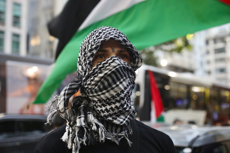 Um manifestante, cujo rosto está envolto num lenço keffiyeh preto e branco, junta-se a um protesto em frente a uma bandeira palestiniana.