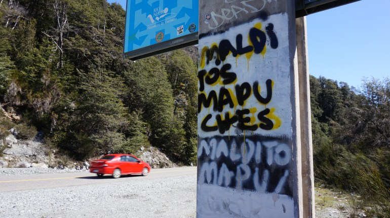 A red car drives by a road sign covered in graffiti, which reads: "Maldi tos Mapuches" or "Damn all Mapuches".