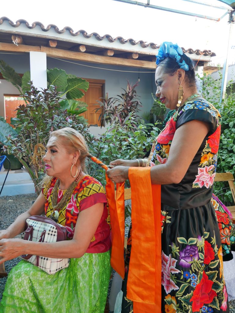 A woman in a blue headdress and a black traditional dress, decorated with flowers, braids another person's hair in the outdoor courtyard of a building, decorated with small palms.
