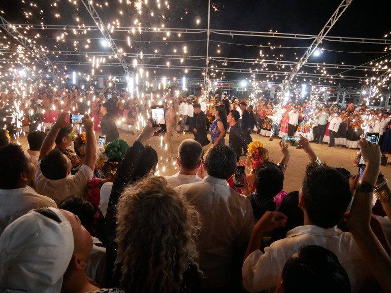 A crowd gathers as fireworks send up golden sparks under a metal-framed structure.