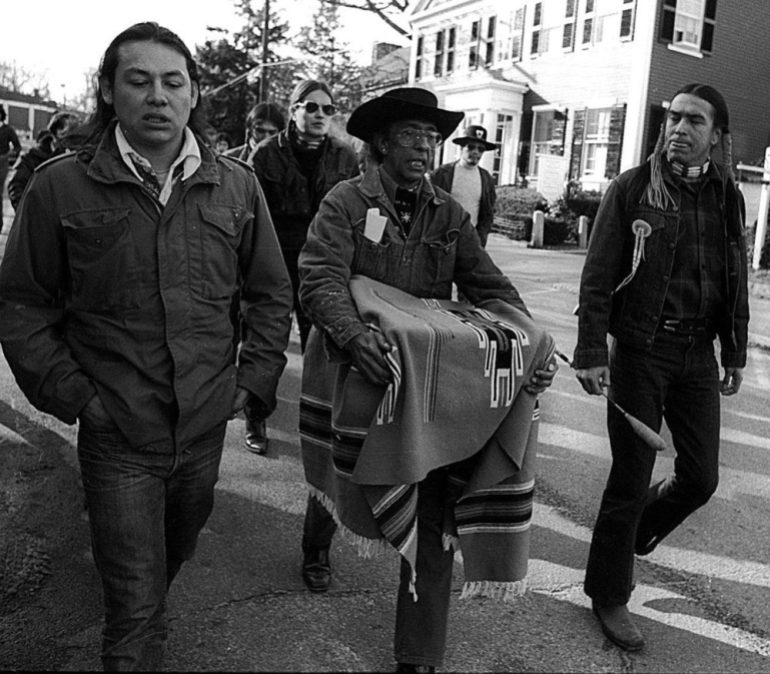 A black-and-white archival photo shows Wamsutta Frank James in the centre of a march holding an Indigenous blanket, as two of his fellow activists walk beside him.