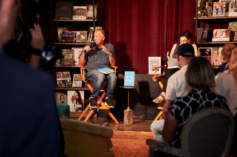 Jeremy Nobel sits in a wooden folding chair, speaking into a microphone on a small wooden stage, where he is surrounded by books. On a small table is his book, Project UnLonely.