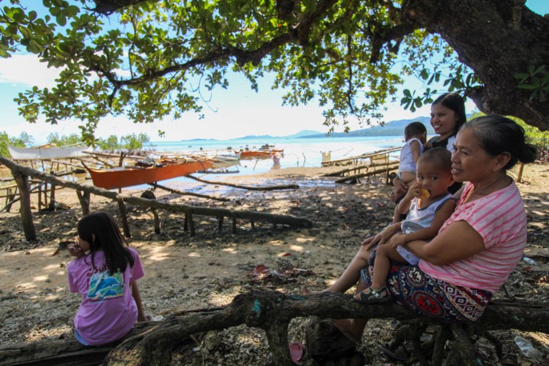 Jecel Espina-Pedel sentada con su familia junto al mar.  Ella está parada y sosteniendo a un bebé.  Los demás están sentados sobre un tronco.