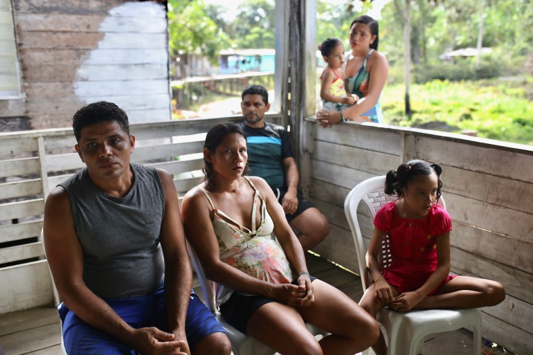Os moradores sentam-se em um deck de madeira, alguns segurando crianças pequenas.  A vegetação da floresta amazônica é visível além do deck.