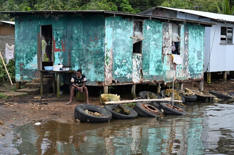 A man sitting outside a wooden house. There is water lapping at the stilts supporting the building and tyres on the ground holding back the water.