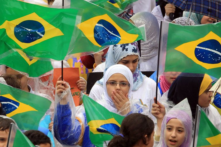 Children in hijabs wave Brazilian flags in an outdoor crowd. One girl covers her mouth with her hand.