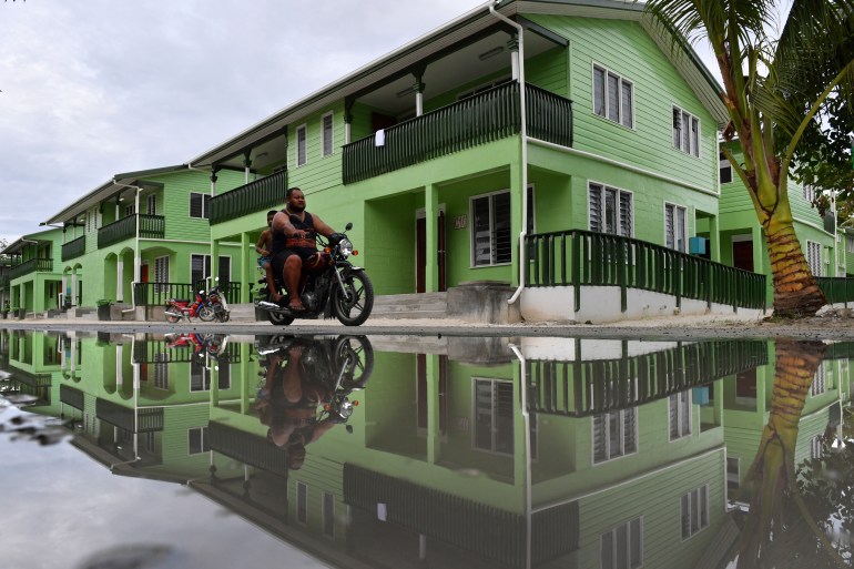 Un hombre que conduce una motocicleta se ve reflejado en un charco de agua en Funafuti, Tuvalu. Los edificios detrás están pintados de verde y tienen balcones.