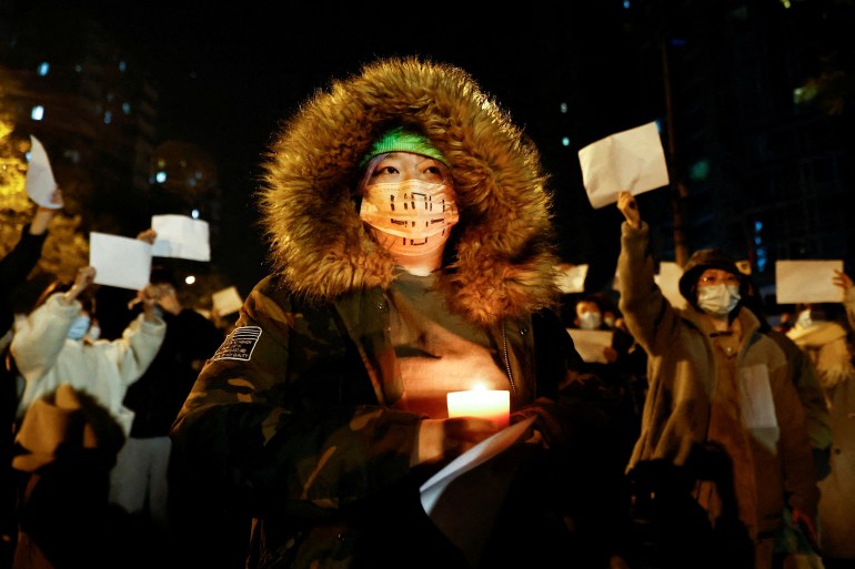 A woman holding a candle during a vigil for people who died in a fire in Urumqi when the building was sealed off because of COVID-19. She's wearing a winter coat with a fluffy hood. Other people are also protesting. 