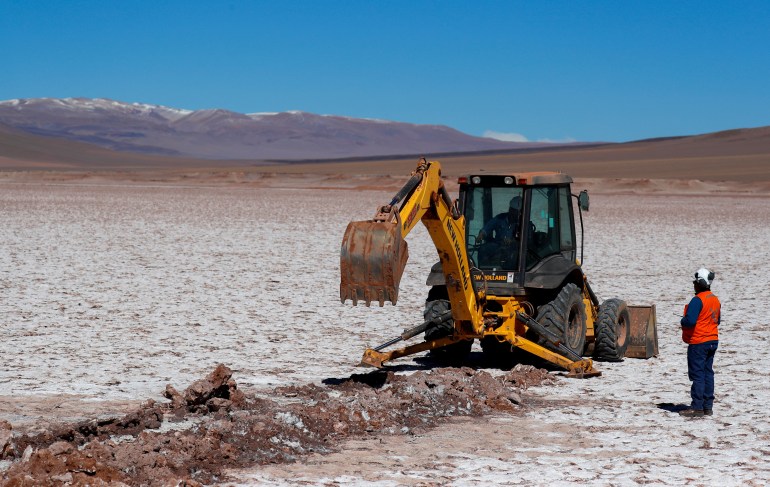 Uma escavadeira industrial – uma máquina com uma pá na frente – está parada em uma salina na Argentina.  Montanhas cobertas de neve podem ser vistas à distância.