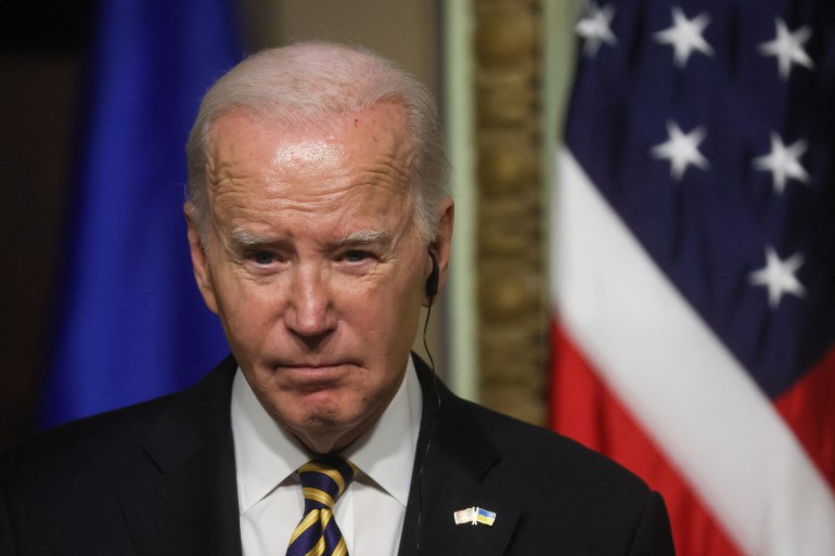 A close-up of Joe Biden, wearing a dark suit and blue-and-yellow patterned tie. He wears an earpiece for translation and a lapel pin showing the Ukraine and US flags. A US flag is visible behind him.