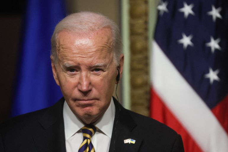 Um close de Joe Biden, vestindo terno escuro e gravata estampada em preto e amarelo.  Uma bandeira dos EUA é visível atrás dele.