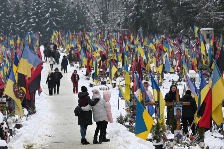 People in a cemetery to pay respects to Ukrainian soldiers killed in the war with Russia. There is snow on the ground. The graves are decorated with Ukrainian flags.