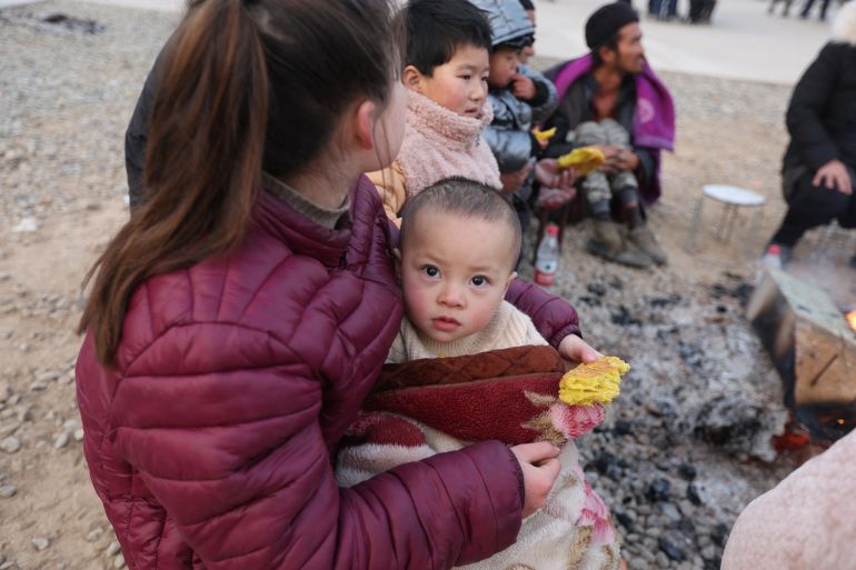 A woman hugs her baby who is wrapped in a blanket as they huddle together in the cold following the earthquake. They are sitting on stools around a fire with other residents.