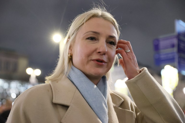 Yekaterina Duntsova. She is standing outside and wearing a cream-coloured coat with a pale blue scarf, She has long blonde hair. 