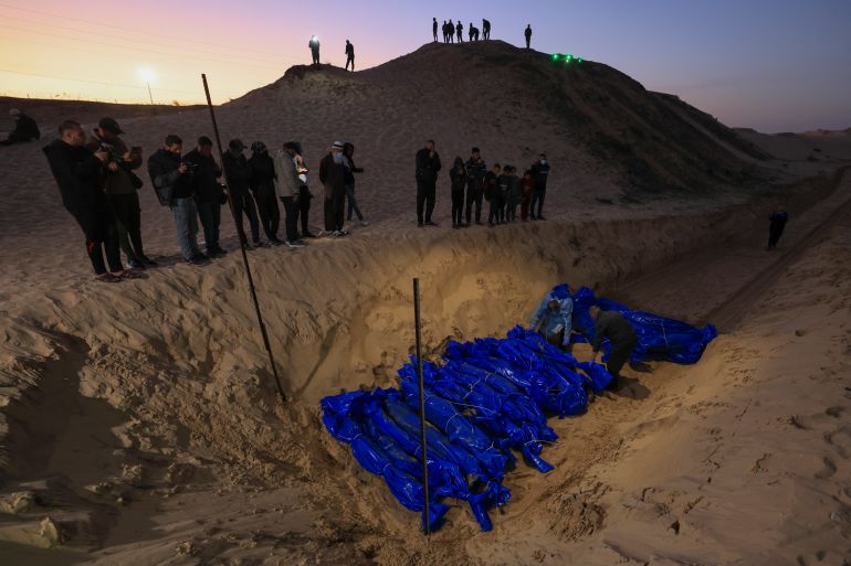 People look on as the shrouded bodies of Palestinians killed in nothern Gaza, that were taken and later released by Israel, are burried in a mass grave in Rafah, on the southern Gaza Strip on December 26, 2023, amid ongoing battles between Israel and the militant group Hamas. (Photo by Mahmud HAMS / AFP)
