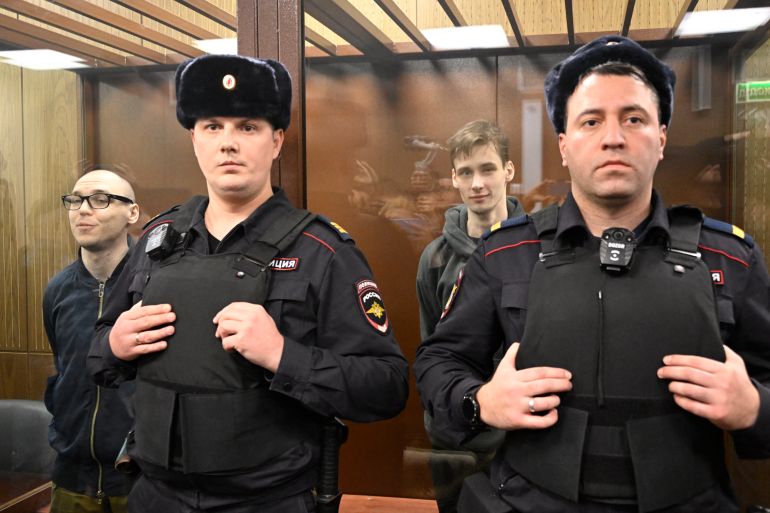 Russian poets Artyom Kamardin (L) and Yegor Shtovba (R) stand inside the defendants' glass cage. There are two police officers in black uniforms and bulletproof vests standing in front of the cage.