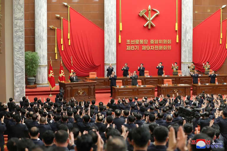 İşçi Partisi toplantısından bir görüntü.  Kim Jong Un podyumun solunda, salondaki delegelerin yanı sıra sahnedeki diğer kişiler tarafından da alkışlanıyor. Arkasındaki duvar kırmızı bayraklar, parti bayrakları ve çekiç ve orakla süslenmiş.