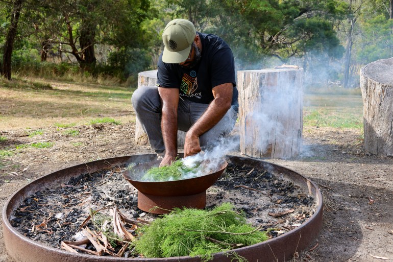 A man conducting a traditional Aboriginal smoking ceremony. He is sqautting down next to a large pan with a smaller one inside. There is greenery in the smaller and smoke rising.