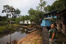 A woman stands on a walkway made of wooden planks that runs along a dried-up river. Houses on stilts sit along the river bank.