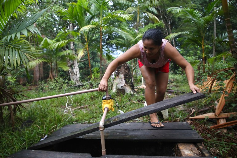 A woman leans over to lift wooden planks from above a well in the Amazon rainforest.