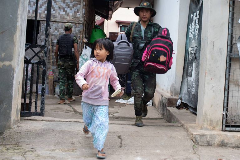 A child running out of a building as she is evacuated. A man behind her is carrying two small backpacks. Another is waiting at the door in the background for more civilians